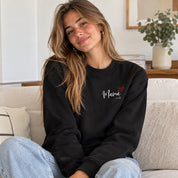 Sudadera negra personalizada con "Mamá", corazón rojo e iniciales de los hijos bordados en el pecho, mujer sentada en el sofá